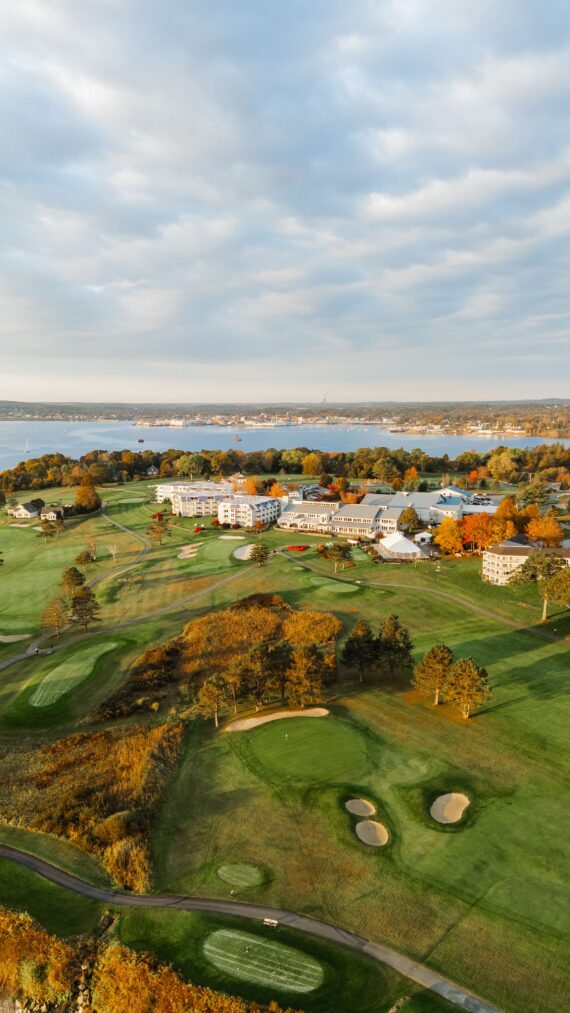 Aerial view of a golf course surrounded by autumn trees. The landscape includes several green fairways, sand bunkers, and a cluster of buildings. A large body of water and a distant town are visible under a partly cloudy sky.
