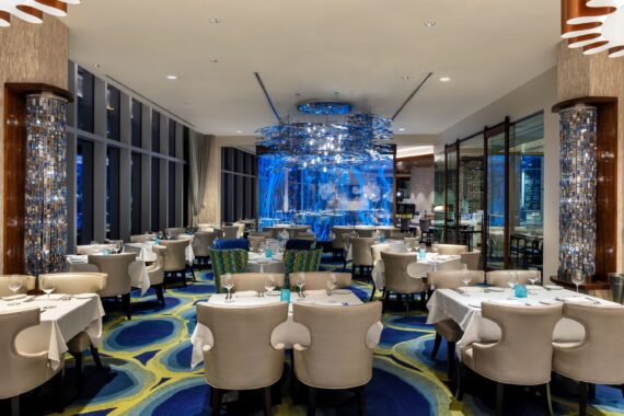 Luxurious restaurant interior with elegant white tablecloths and plush chairs arranged neatly around tables. A vibrant blue aquarium feature and modern ceiling design add an upscale ambiance to the space. Large windows provide natural light.