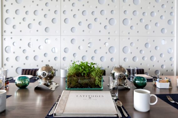 A modern dining table setup with a Latitudes menu, white coffee cup, small potted plants, and decorative silver domes. The backdrop features a white wall with circular patterns, enhancing the contemporary ambiance.
