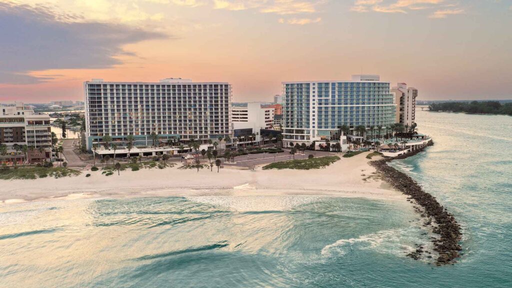 Aerial view of two new hotels in Clearwater Beach, Florida, featuring large windows on a sandy shore lined with palm trees and a rocky jetty stretching into the turquoise ocean beneath a pastel sunset sky.