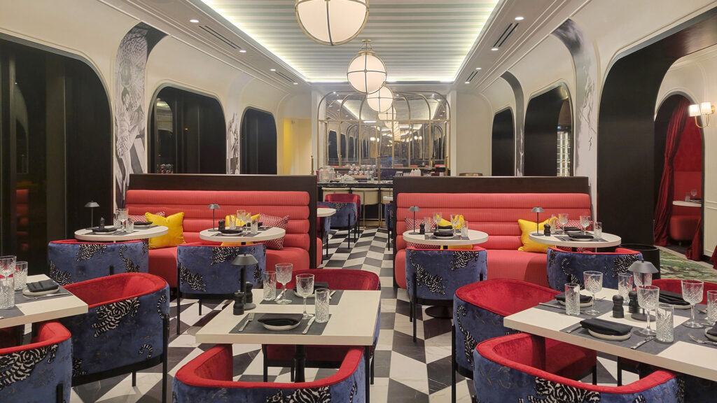 The stylish restaurant, Ringside at Cirque, with an interior with checkerboard floors, red and blue upholstered chairs, pink cushioned booths, round white tables set for dining, and soft overhead lighting.