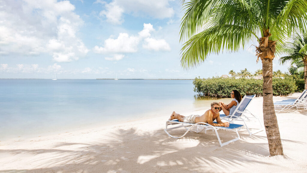 Two people relax on lounge chairs under a palm tree on a sandy beach. The calm sea stretches out to the horizon under a bright blue sky with scattered clouds.