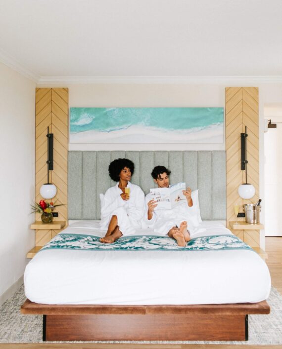 A couple in white robes relax on a neatly made bed in a modern bedroom. They are looking at a magazine, with a large wall art of an ocean scene above the bed. The room has wooden accents, a vase with flowers, and a bucket beside the bed.