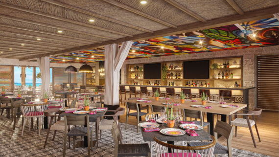 Spacious restaurant with colorful mosaic ceiling and woven wood decor. Tables are set with pink and white dishes. A well-stocked bar with shelves of bottles is in the background, with wooden chairs and decorative lights. Large windows offer an ocean view.