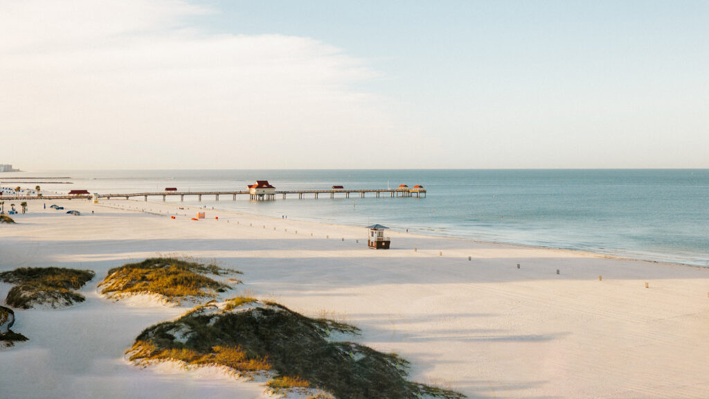 A serene beach scene unfolds in Clearwater, Florida, with a long pier extending into calm, blue waters. Sandy dunes with patches of grass grace the foreground. The clear sky holds a whisper of clouds near the horizon, while small structures dot this idyllic attraction on the pier.