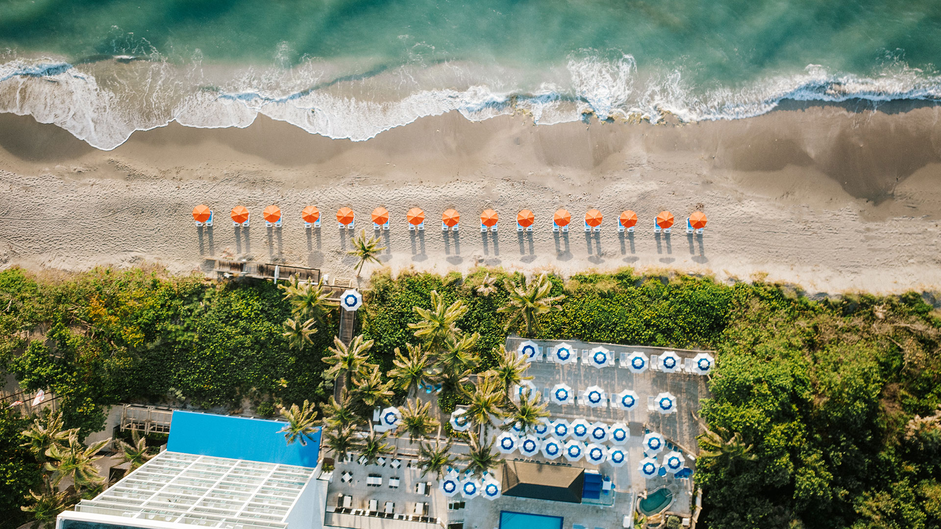 Aerial view of a beach with waves washing onto the shore. Orange umbrellas line the sandy beach. Below the umbrellas, a resort pool area with blue and white circular sunshades is visible, surrounded by palm trees and greenery.