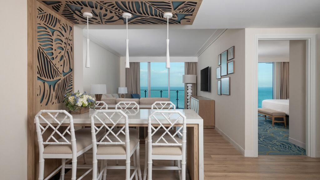 Modern hotel suite with a dining table and four white chairs, a living area with a sofa, large windows showing an ocean view, and a separate bedroom visible through an open doorway. Decorative patterns adorn the ceiling and wall.
