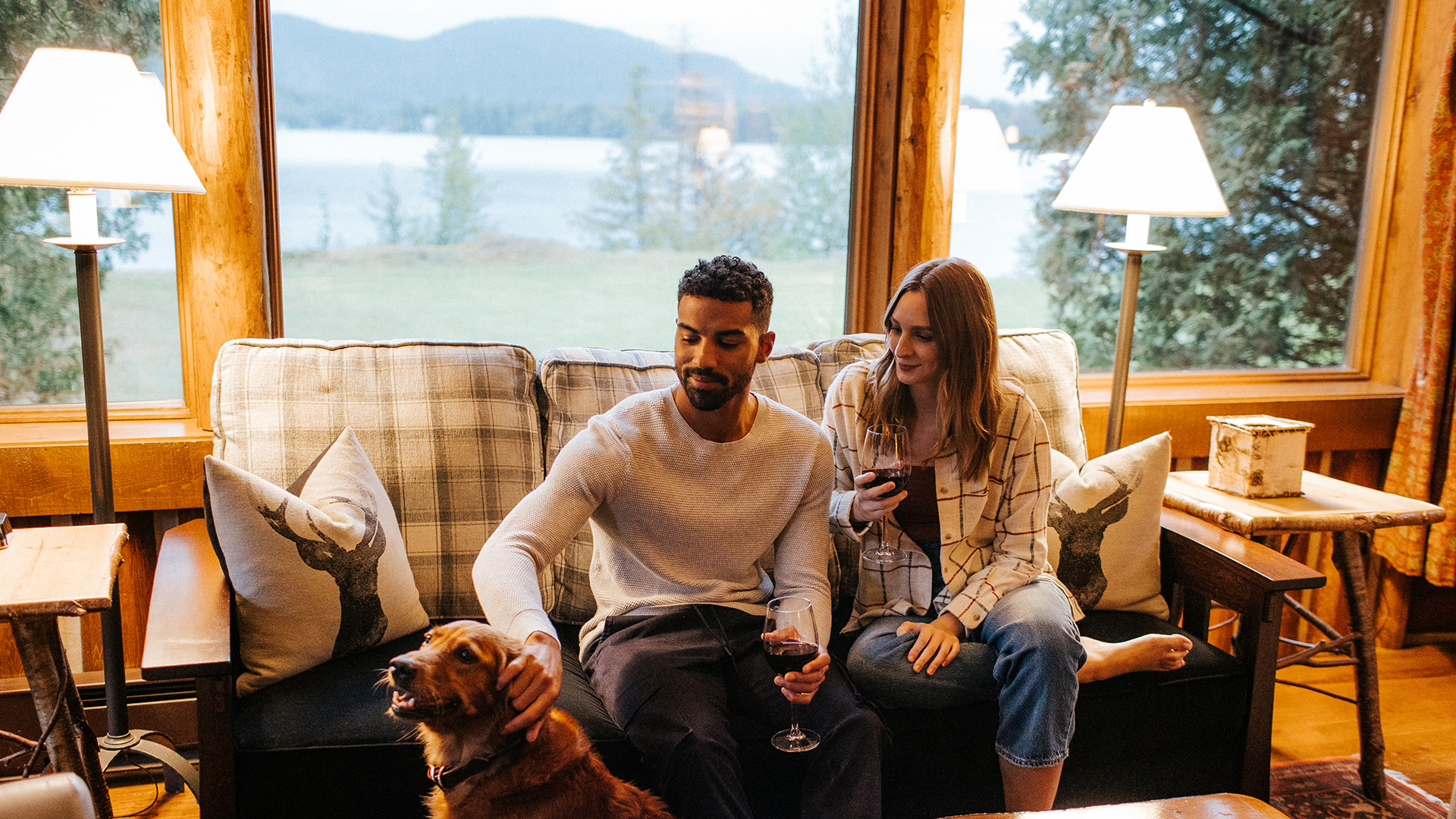 A man and woman sit on a cozy couch in a cabin, holding glasses of red wine. The man pets a brown dog. Large windows behind them reveal trees and a lake with mountains in the distance. Warm lamps light the room.
