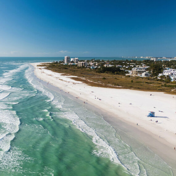 Aerial view of a wide sandy beach with turquoise waves, a few people walking along the shore, and buildings and greenery in the background under a clear blue sky.