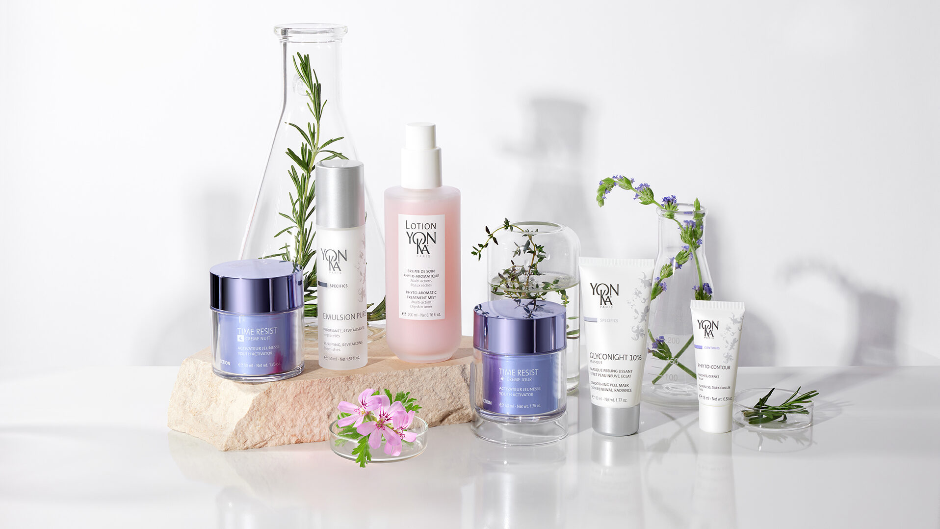 Six Yon-Ka skincare products are arranged on a white surface with flowers, small glass bottles, and sprigs of greenery for decoration. The products include jars, bottles, and tubes in soft pastel colors.