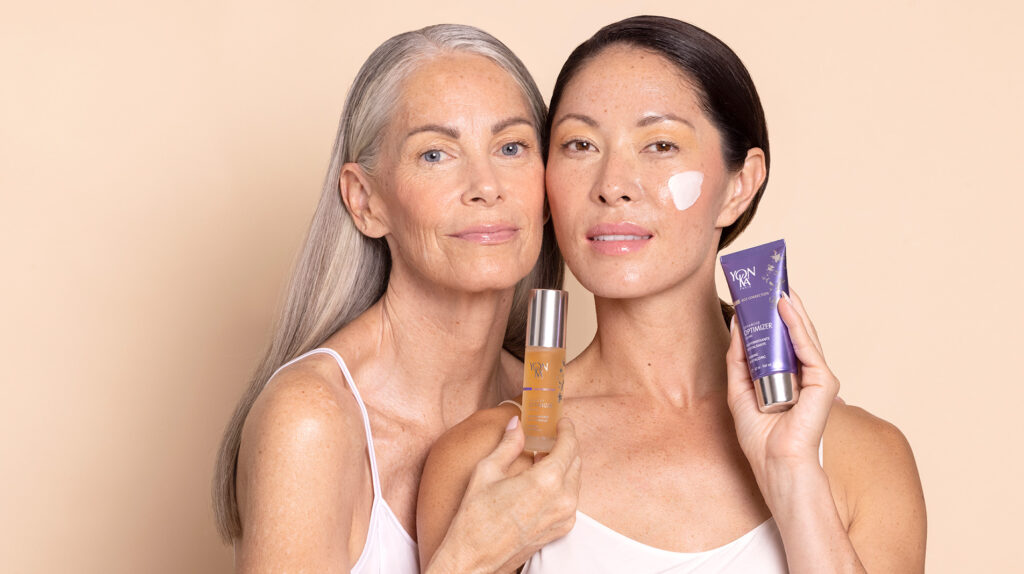 Two women with clear skin, one older with gray hair and one younger with dark hair, smile softly. The older woman holds a serum bottle, while the younger has cream on her cheek and holds a purple skincare tube.