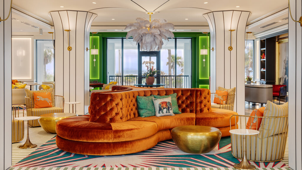 A stylish hotel lounge at Hotels On Lido Beach Sarasota features a curved orange velvet sofa, gold tables, colorful pillows, a feathered chandelier, and large windows overlooking palm trees and a balcony. The decor boasts vibrant colors and modern eclectic touches.