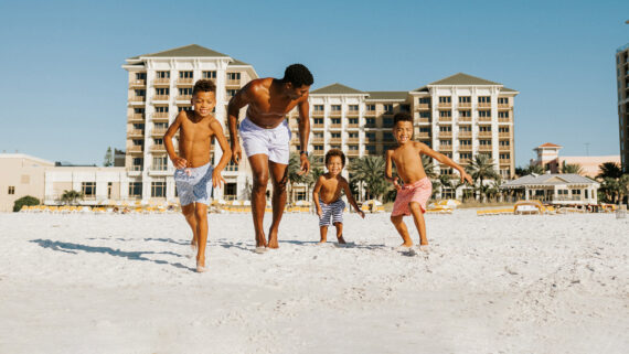 A man and three children, all wearing swimwear, run and play together on a sandy beach with Sandpearl Resort in the background.