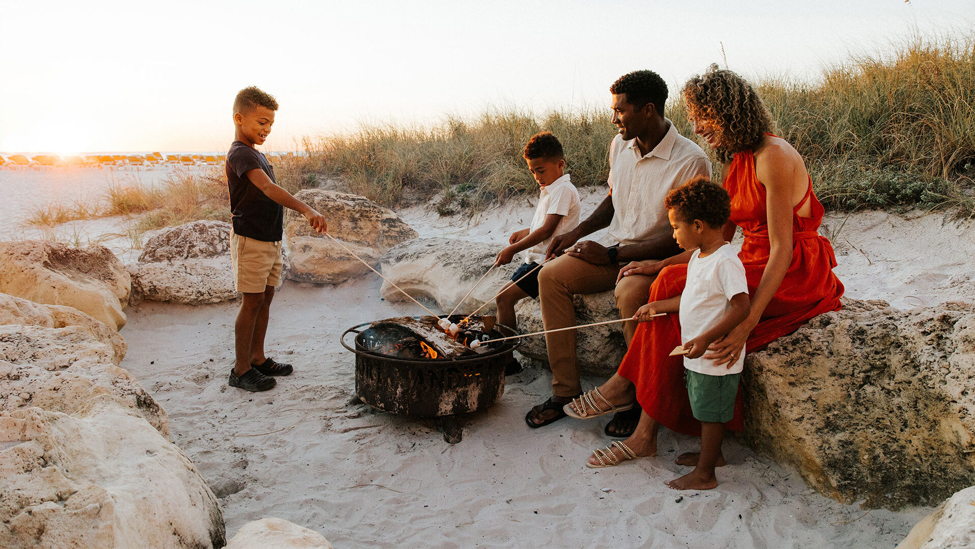 A family of five sits on rocks at the beach, roasting marshmallows over a fire pit at sunset. The children and parents are smiling and enjoying the warm, sandy setting together.