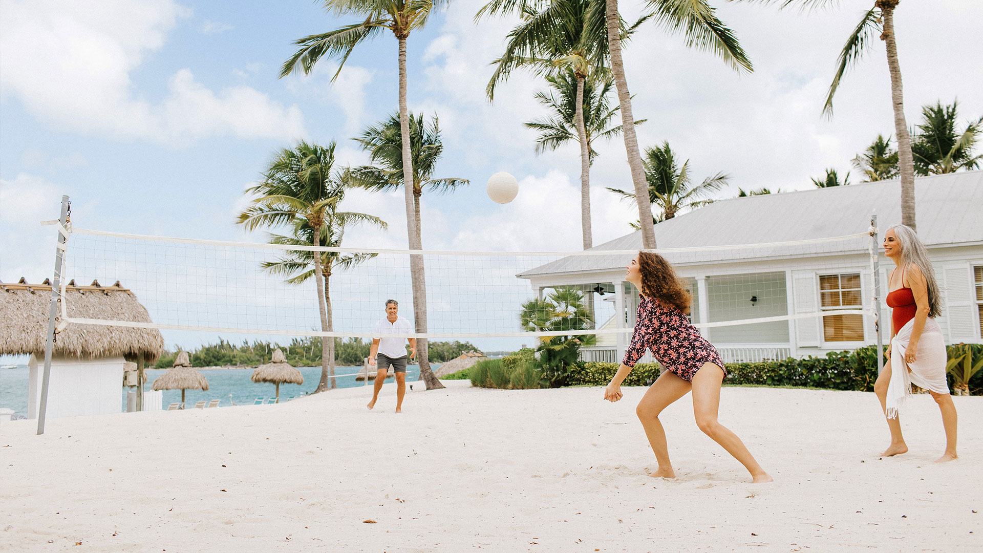 Three people play beach volleyball on white sand surrounded by palm trees, with tropical huts, ocean, and a white house in the background under a partly cloudy sky.