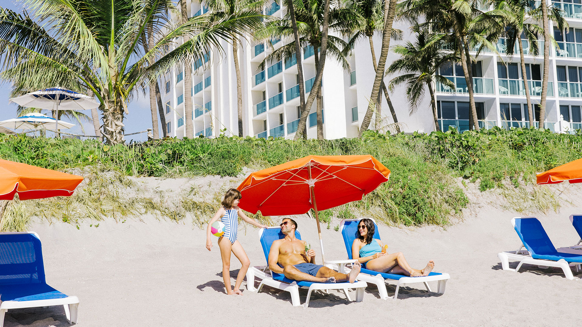 A family relaxes on a sandy beach under a red umbrella, with a man and woman sitting on lounge chairs and a young girl standing nearby. Palm trees and Jupiter Beach Resort are in the background.