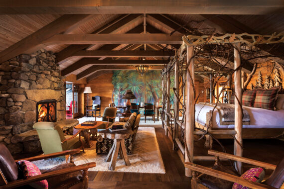A cozy cabin interior with wooden beams, a canopy bed, and stone fireplace. The room is warmly lit, featuring rustic furniture, a large mural on the back wall, and a dining area set by the window.