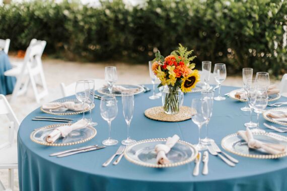 An elegantly set round table with a teal cloth, featuring plates and cutlery arranged neatly. A floral centerpiece with sunflowers and other blooms adorns the table. Crystal glassware surrounds each setting. Greenery is seen in the background.