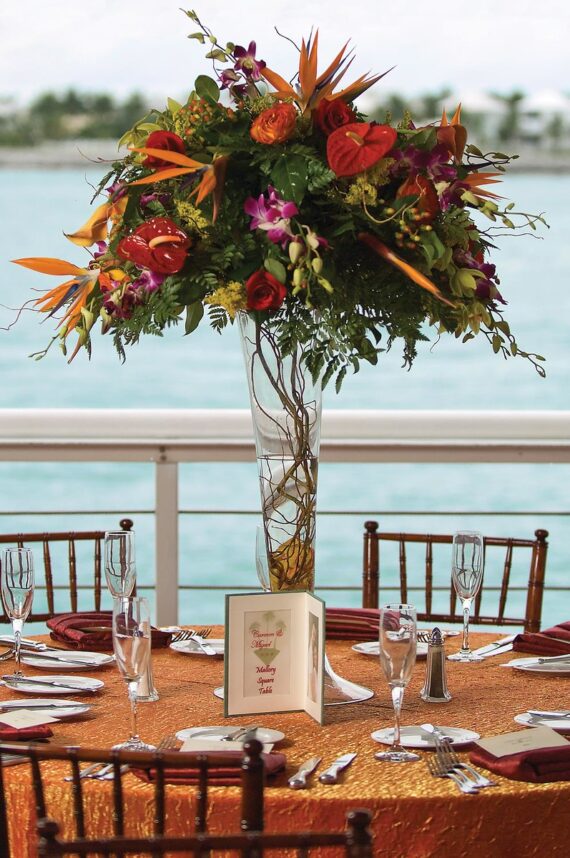 A tall floral centerpiece with vibrant flowers and greenery sits atop a round table covered in an orange cloth. The elegant setup features wine glasses, menus, and wooden chairs. The background shows a view of water and distant trees.