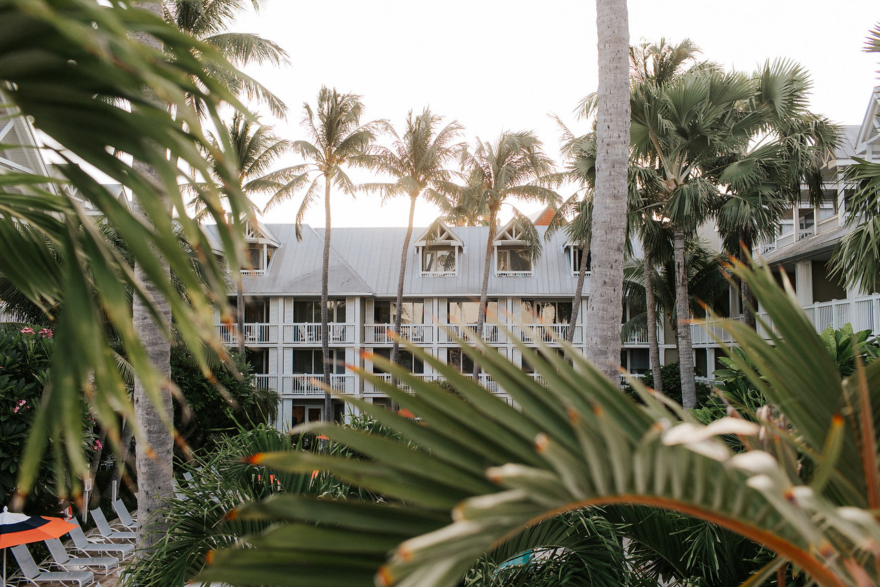 A resort building surrounded by tall palm trees and lush greenery. The structure features multiple balconies and an A-frame roof. The sun is setting behind the building, casting a warm glow over the scene.
