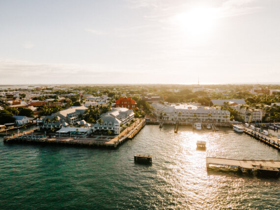 Aerial view of a coastal city with a pier extending into the water. Sunlight reflects on the ocean, and buildings with white and red roofs line the waterfront. The sky is clear with the sun low on the horizon.