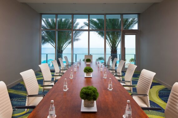 A modern conference room with a long wooden table surrounded by white chairs. The table is set with water bottles and small potted plants. Large windows offer a view of palm trees and the ocean, providing a glimpse of Clearwater Beach in the background.