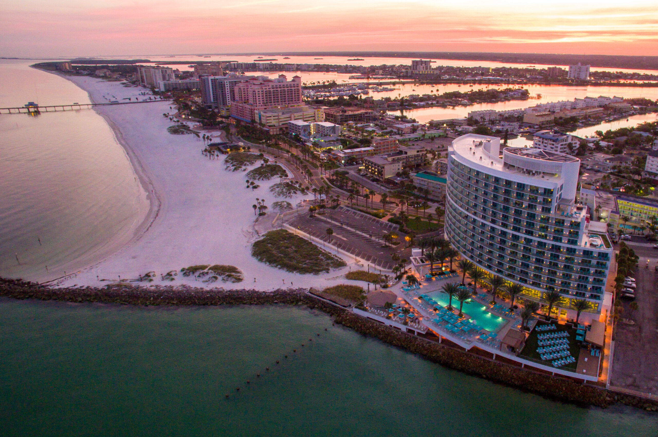 Aerial view of a beachfront city at sunset. A curved modern hotel with a pool is in the foreground, overlooking a sandy beach. The cityscape stretches along the coast with a mix of buildings and a pier extending into the water.