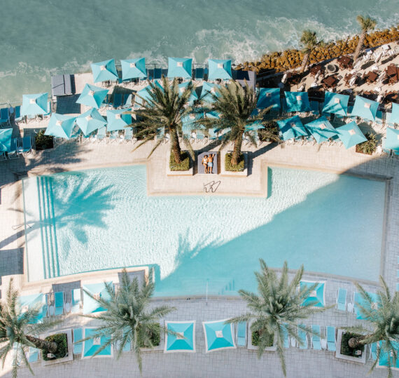 Aerial view of a resort pool at Clearwater Beach, surrounded by palm trees and turquoise sun loungers. The poolside area overlooks the ocean, with waves gently crashing on rocks. Two people are sunbathing on loungers under an umbrella near the pool's center.
