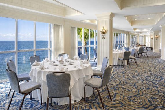 Elegant dining area with round tables set for a formal event. Large windows offer a scenic ocean view, with palm trees visible outside. The room features patterned carpet and decorative lighting, providing a bright and sophisticated atmosphere.