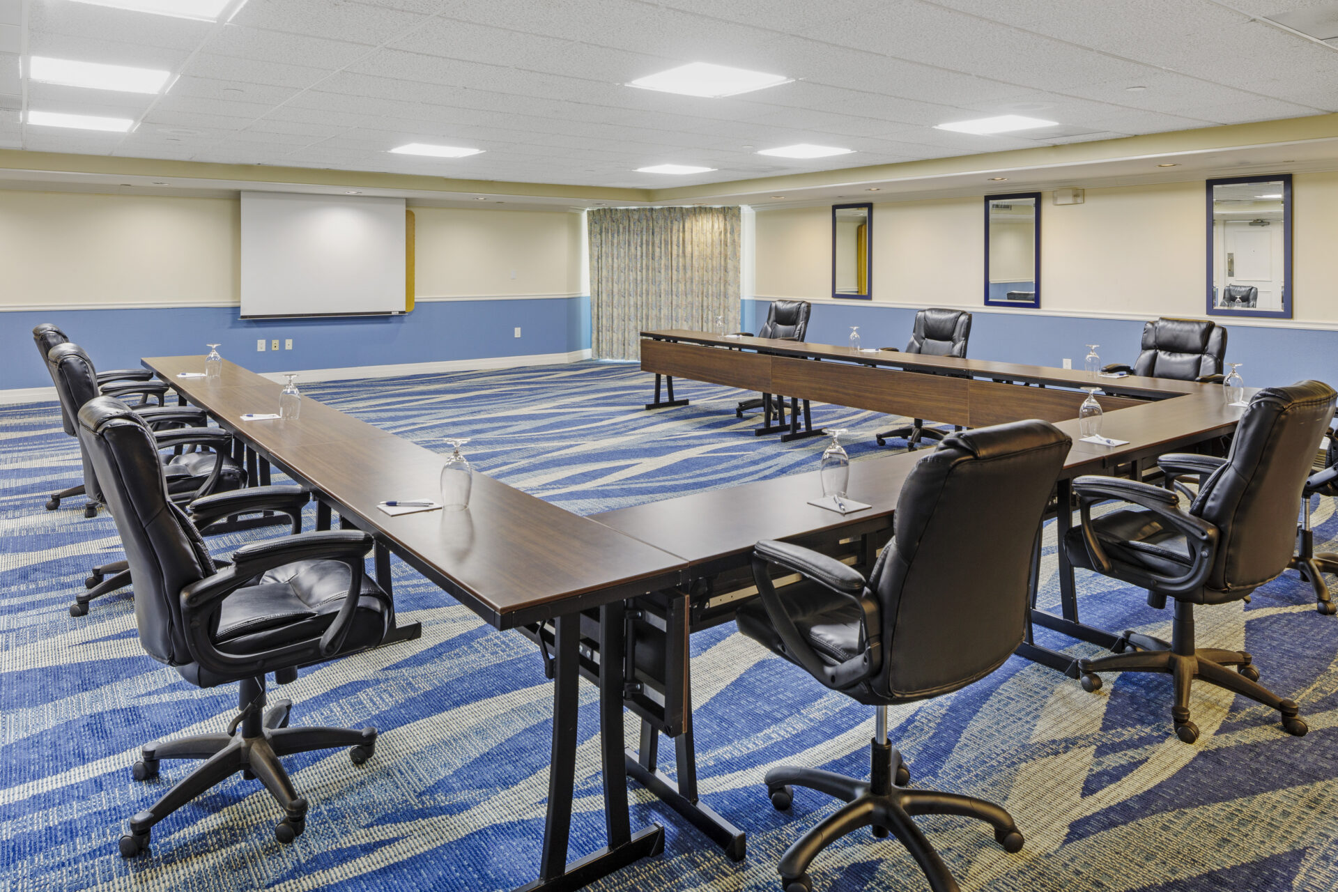 A conference room with U-shaped table arrangement, equipped with black leather office chairs. There are water glasses on the tables, a projector screen on one wall, and patterned blue carpeting on the floor. The room is well-lit.