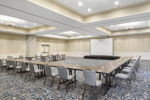 A spacious, empty conference room with U-shaped table setup. Chairs are arranged around the tables, and a large projector screen stands at the front. The room has patterned carpet and elegant light fixtures on the ceiling.