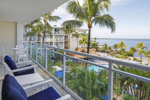 A hotel balcony with white furniture overlooks a pool area surrounded by palm trees. Beyond the pool, theres a view of the ocean and a partly cloudy sky.