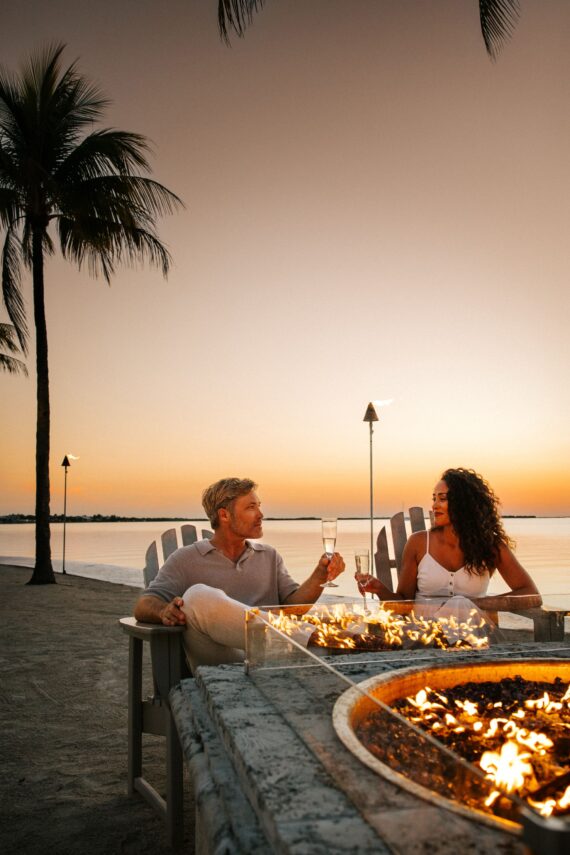 A couple sits by a beachside fire pit, toasting with champagne glasses at sunset. They are surrounded by palm trees, with the ocean in the background. The sky is a warm gradient of colors as day transitions to night.