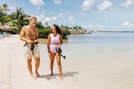 A couple walks along the beach with snorkeling gear in hand.
