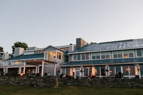 A large, modern building with a metal roof is set against an evening sky. Warm lights glow from the windows, and people are seated on a patio with a stone wall in the foreground.