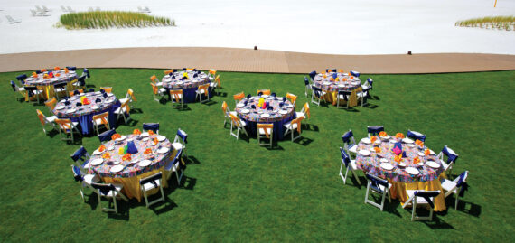 Outdoor dining setup on a grassy lawn with several round tables arranged for an event. Each table is adorned with colorful tablecloths, place settings, and centerpieces. A sandy area and boardwalk are visible in the background.