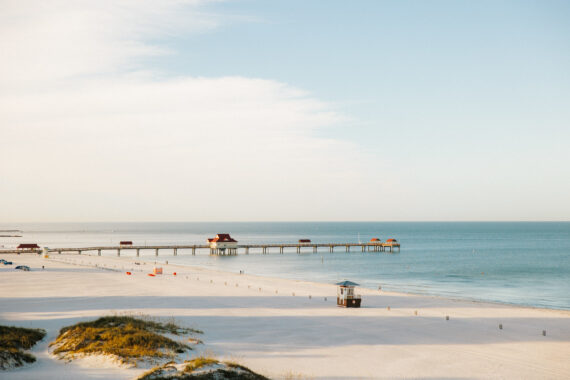 A serene beach scene with a long pier extending into the calm blue ocean. The sandy shore is dotted with patches of grass, and the sky is clear with a few clouds. A small lifeguard tower is visible in the foreground.