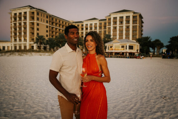 A couple stands on a sandy beach at sunset, with a large hotel in the background. They smile while holding glasses of wine, with the woman in a red dress and the man in a cream shirt and brown pants.