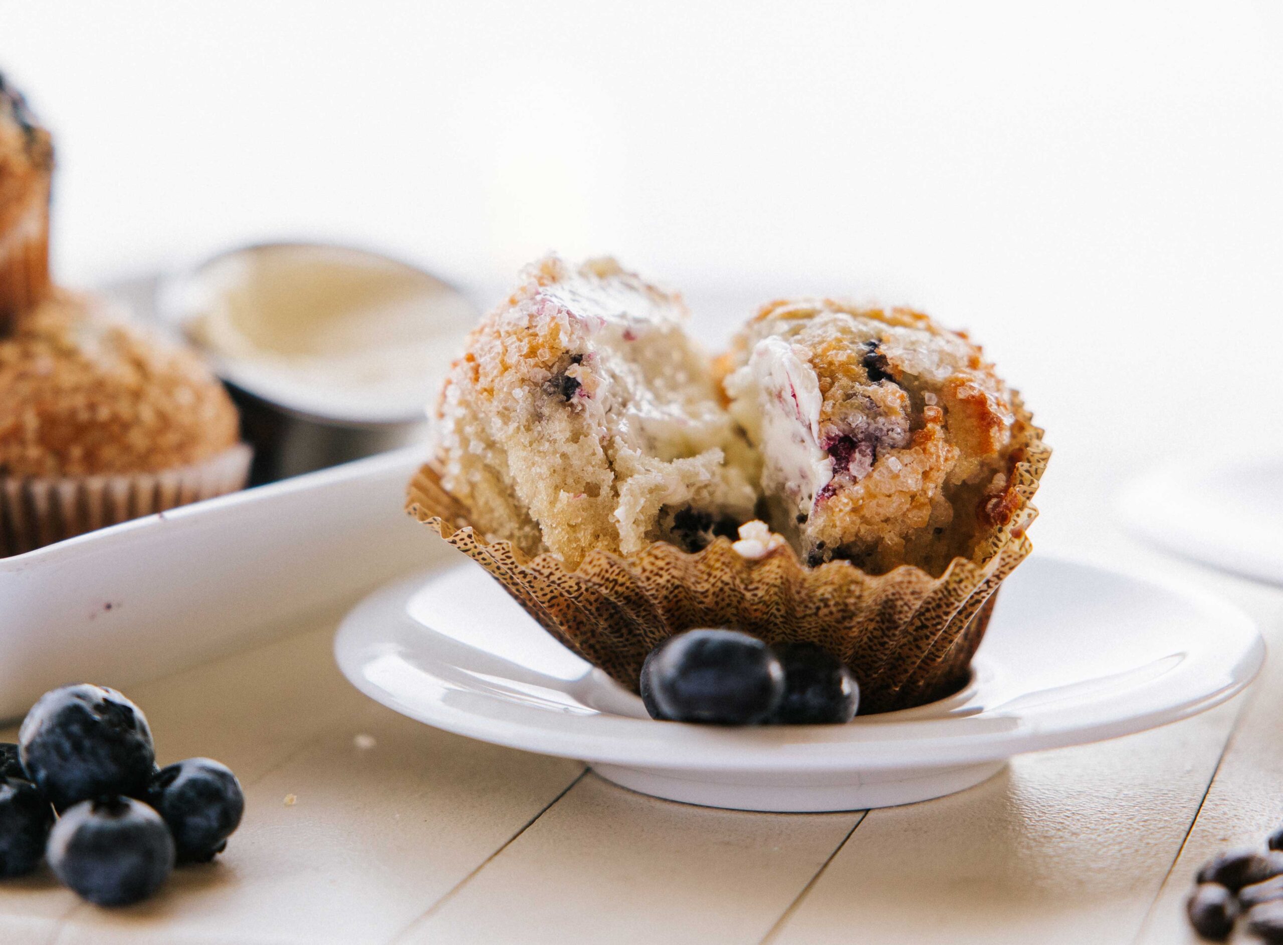 A split blueberry muffin on a white plate, revealing its soft, crumbly interior. Fresh blueberries are scattered around the table, and theres a blurred muffin tin in the background.