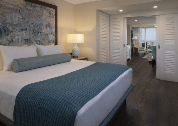 A cozy hotel room featuring a neatly made king-size bed with teal bedding, a large artwork above the headboard, and a bedside lamp. In the background, there is a seating area with a view of a balcony and the ocean.