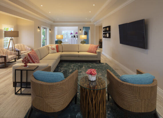 A spacious living room with a large L-shaped sofa, two armchairs with blue cushions, and a round glass coffee table. A flat-screen TV is mounted on the wall and there are decorative lamps on a console table near the window.