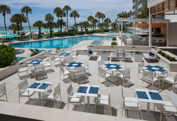 Outdoor dining area overlooking a pool with white tables and chairs, and blue placemats. Palm trees and the ocean are visible in the background, under a clear blue sky.