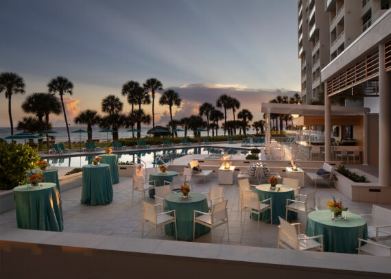 An outdoor party setup at a resort with tables covered in teal cloths and floral centerpieces. Theres seating around fire pits, a pool nearby, and palm trees silhouetted against a sunset sky. A tall building is on one side.