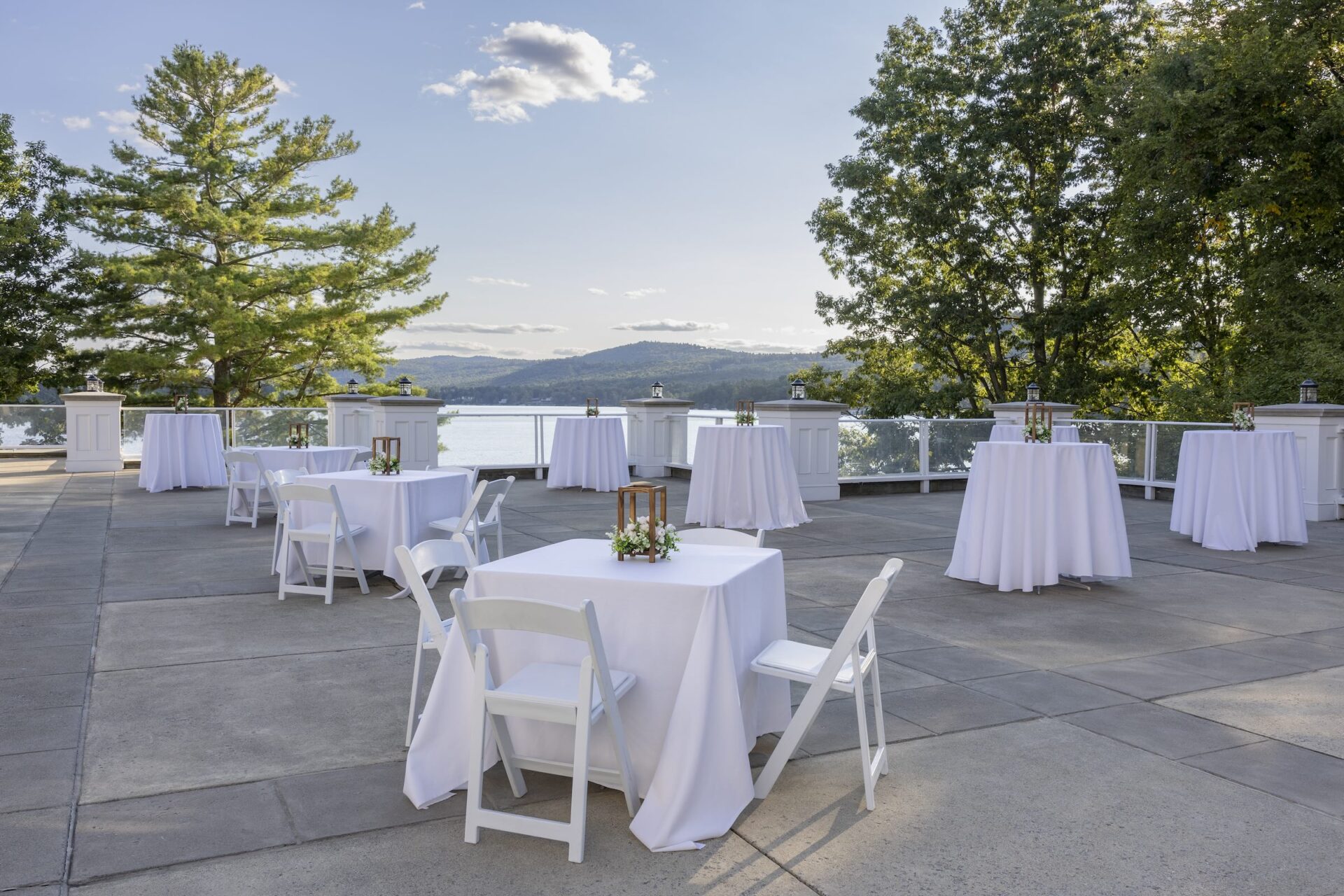 An outdoor patio set for an event with several round and square tables covered in white tablecloths. Each table has floral centerpieces. The background features a lake, distant hills, and trees under a clear sky.