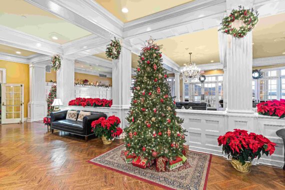 A festive living room is adorned with a Christmas tree decorated with red and gold ornaments. Wreaths and poinsettias accent the space, which features white columned walls, a dark sofa, and a chandelier, creating a warm holiday atmosphere.