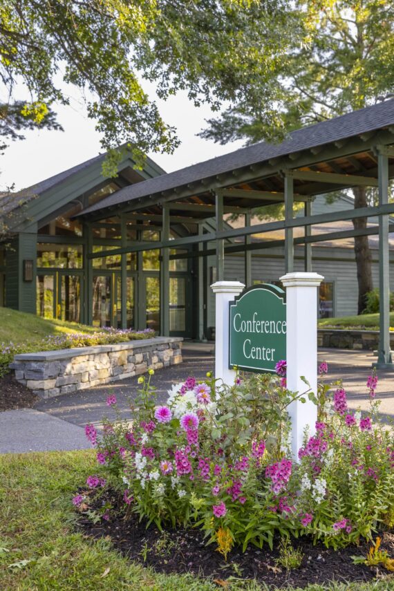 A green building with large windows and a gabled roof is surrounded by trees and flowers. In the foreground, a white sign with green trim reads Conference Center, framed by colorful blossoms.