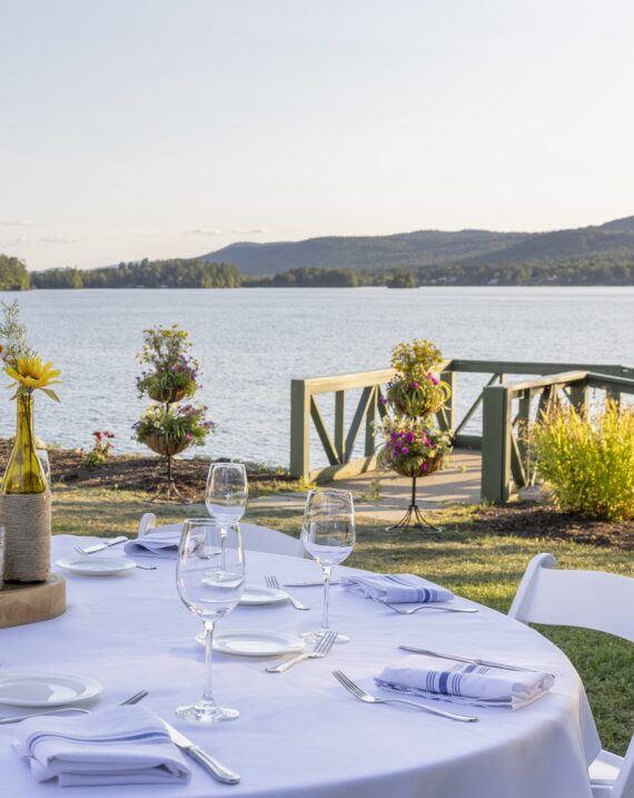 A round table with a white tablecloth is set with glassware and napkins beside a lake. Decorative flower arrangements and a wooden deck overlook the water. Hills and trees are visible in the distance under a clear sky.