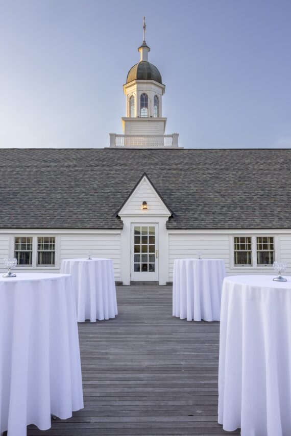 A rooftop setting with circular tables covered in white cloths. In the background, theres a white building with a steep roof and a cupola topped with a spire, under a clear blue sky.