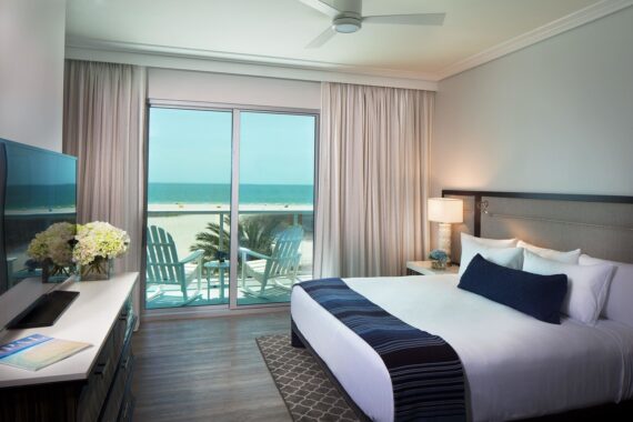A bright hotel room with a king-sized bed, white and blue bedding, and a large window leading to a balcony. Outside, theres a view of a sandy beach and blue ocean. The room features a large TV, flowers, and modern decor.