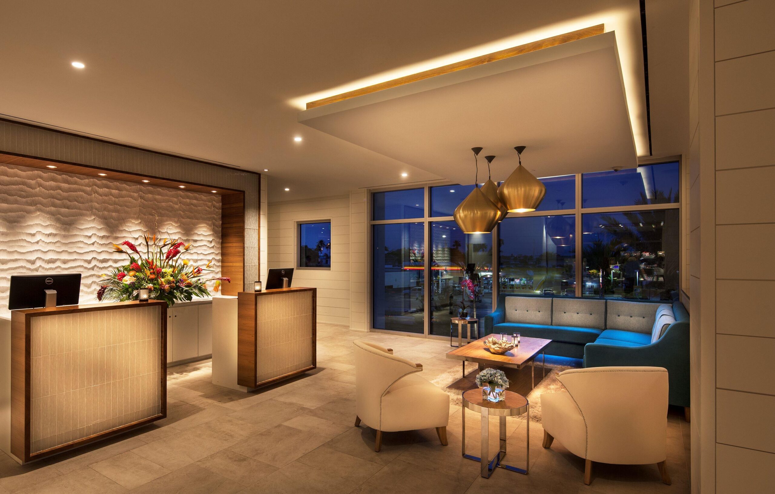 A modern hotel lobby with elegant lighting. The reception desk is decorated with flowers. Comfortable seating includes white armchairs and a blue sofa. Large windows reveal a nighttime cityscape. Gold pendant lights add a touch of luxury.